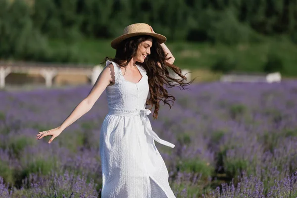 Happy brunette woman in white dress and straw hat walking in field with blossoming lavender — Photo de stock