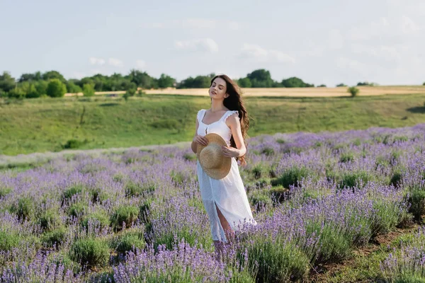 Brunette woman in white dress standing with closed eyes and holding straw hat in lavender field — Foto stock