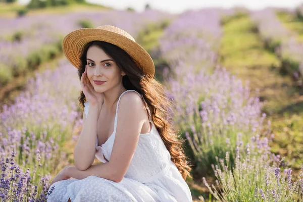 Pretty woman in straw hat and white dress smiling at camera in blurred field with blossoming lavender — Stock Photo