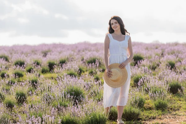 Happy woman in summer dress holding straw hat while standing in lavender field — Stockfoto