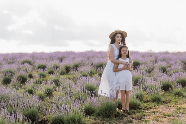 Brunette woman in white dress and straw hat embracing daughter in flowering field - foto de stock