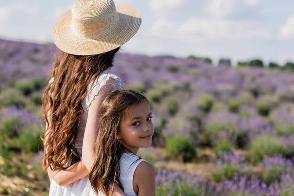 Happy child looking at camera near mom in straw hat in blurred field — Photo de stock