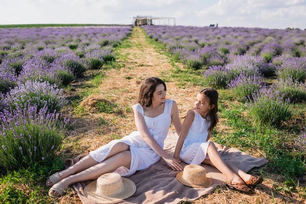 Mother and daughter in white dresses looking at each other near straw hats on blanket in meadow - foto de stock