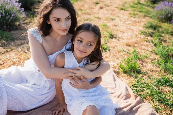 Happy brunette woman embracing daughter while sitting in field on summer day — Fotografia de Stock