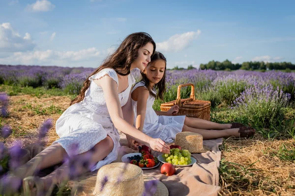 Brunette mother and daughter eating fresh fruits on picnic in meadow — Stockfoto