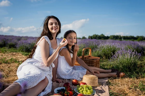 Smiling woman and kid looking at camera while eating fresh fruits on picnic - foto de stock