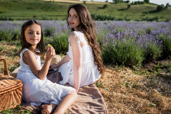 Smiling girl holding grape near mother on picnic in flowering field — Stockfoto