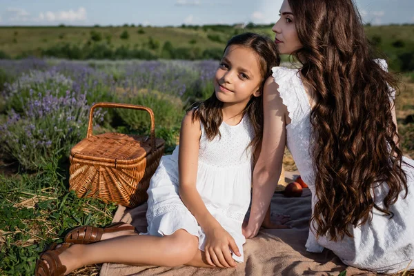 Счастливая девочка смотрит на камеру рядом с мамой и плетеной корзиной в поле — стоковое фото