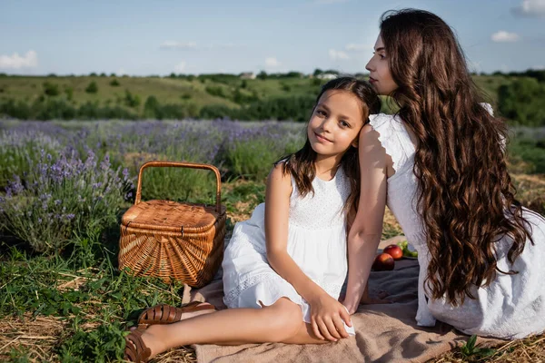 Joyful girl looking at camera near brunette mom and straw basket in lavender meadow - foto de stock