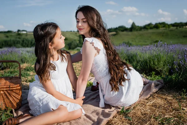 Brunette woman and girl smiling at each other while sitting on blanket in countryside — Foto stock