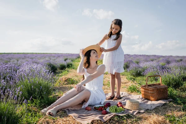 Girl holding straw hat near happy mom sitting on blanket near fresh fruits — Stockfoto