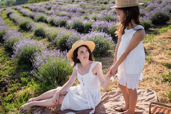 Child in straw hat holding hands of mother sitting on blanket in field — Stock Photo