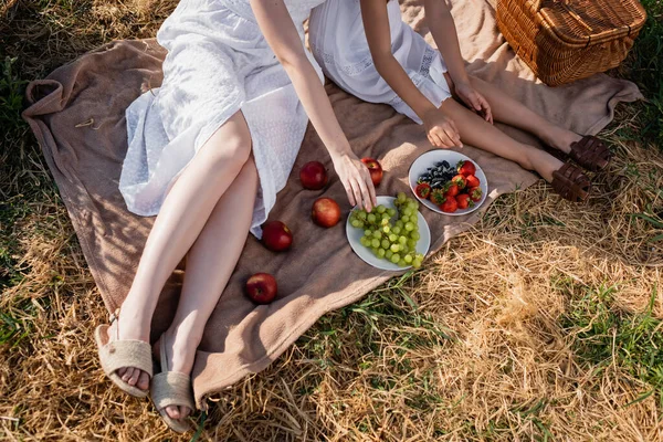 Обрезанный вид женщины и девушки в белых платьях, сидящих рядом со свежими фруктами на одеяле в поле — стоковое фото