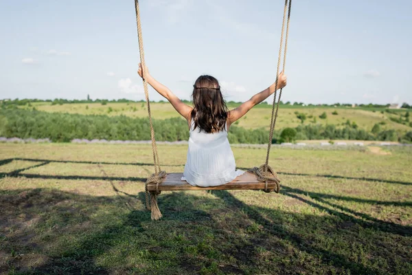 Back view of child in summer dress riding swing in meadow — Fotografia de Stock