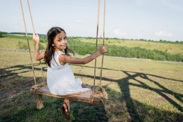 Girl in white dress riding swing and looking at camera in meadow — Stockfoto