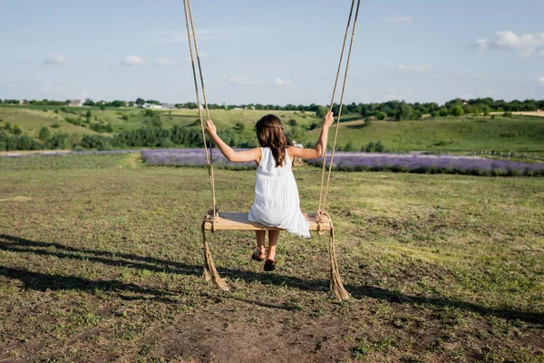 Back view of girl riding swing in field on summer day — Stockfoto