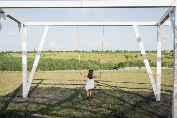 Back view of child in summer dress riding swing in field at countryside — Photo de stock