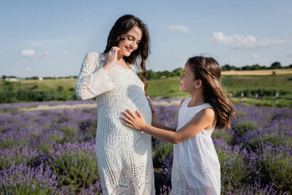 Беременная женщина в белом платье улыбается рядом с дочерью обнимая ее живот на открытом воздухе — стоковое фото