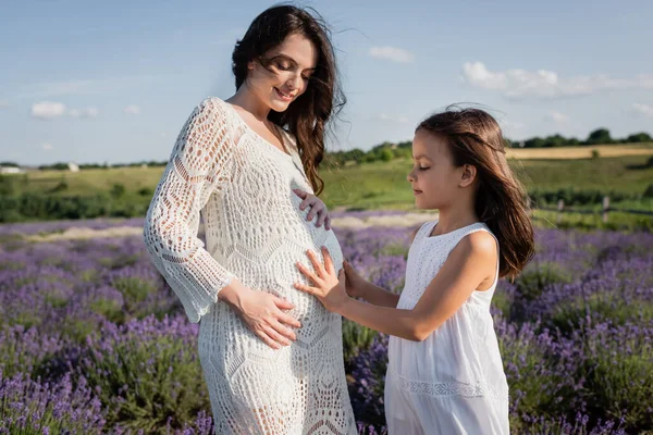 Happy pregnant woman smiling near daughter touching her belly in blurred meadow — Foto stock