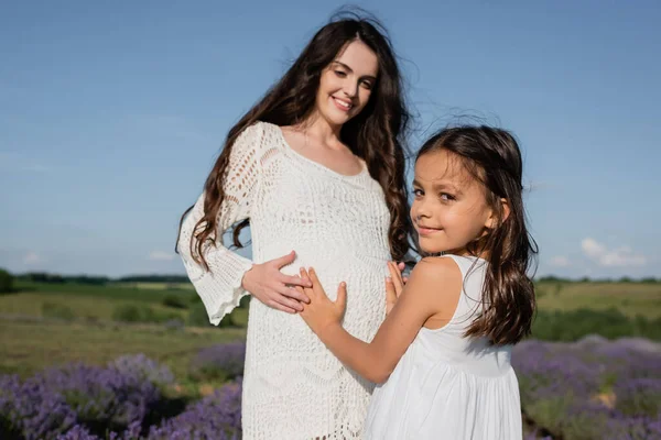 Smiling girl embracing tummy of pregnant mother in openwork dress — Photo de stock