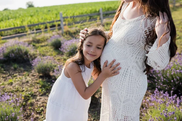 Улыбающийся ребенок обнимает живот беременной матери на размытом загородном поле — стоковое фото