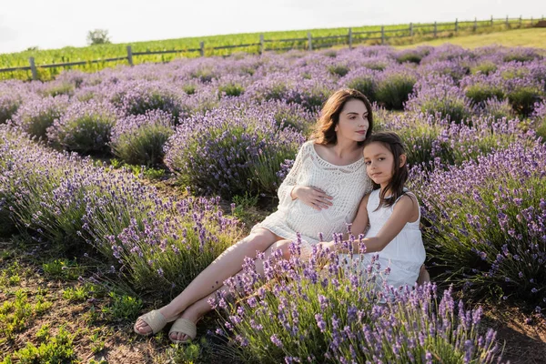 Pregnant woman in white dress sitting with daughter in field with blooming lavender — Photo de stock