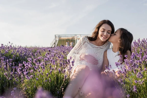 Child kissing pregnant mother sitting in meadow near blooming lavender — Stockfoto