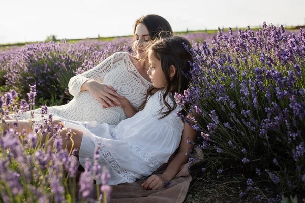 Girl in white dress sitting near mother in field with blossoming lavender - foto de stock