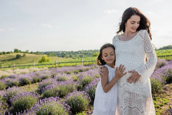 Happy girl embracing tummy of pregnant mother in blooming lavender field — Fotografia de Stock