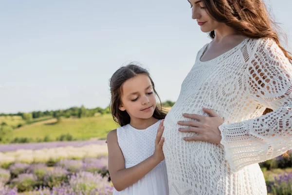 Smiling girl hugging tummy of pregnant mother in blurred field — Photo de stock