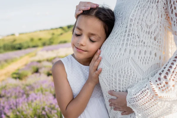 Happy girl with closed eyes leaning on tummy of pregnant mom outdoors — Stockfoto