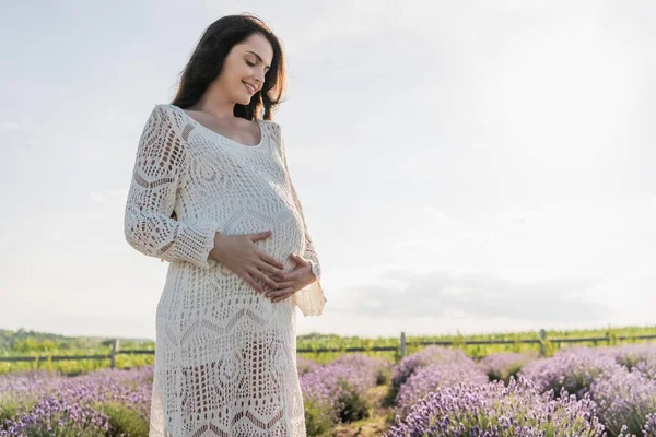 Счастливая беременная женщина в платье касаясь живота в поле с лавандовыми цветами — стоковое фото