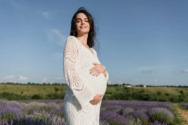 Brunette pregnant woman in openwork dress smiling at camera in lavender field — Stock Photo