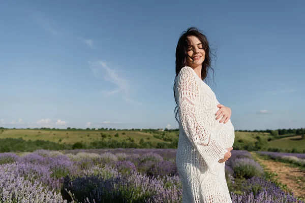 Pregnant woman in white dress standing with closed eyes in lavender field — Fotografia de Stock