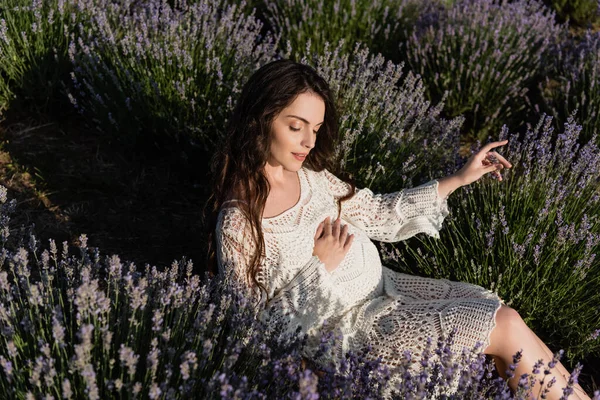 High angle view of pregnant woman with closed eyes touching tummy while sitting in lavender field — Stockfoto