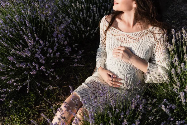 Частичный вид счастливой беременной женщины в платье с открытыми дверями, сидящей на лавандовом лугу — стоковое фото
