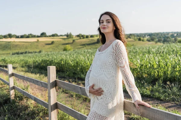 Happy pregnant woman in white openwork dress standing near wooden fence in field — Stockfoto