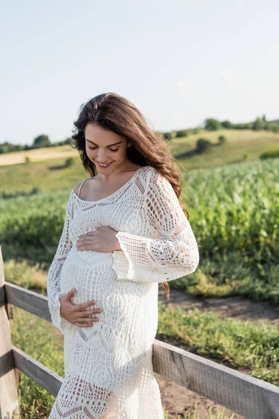 Pretty pregnant woman in white dress touching belly and smiling outdoors — Photo de stock