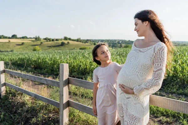 Girl and pregnant woman in dress looking at each other near fence in meadow - foto de stock