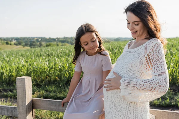 Girl in pink dress sitting on wooden fence near happy and pregnant mother - foto de stock