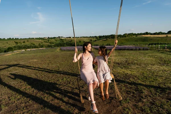 Full length of woman and kid in summer dresses riding swing in meadow — Fotografia de Stock