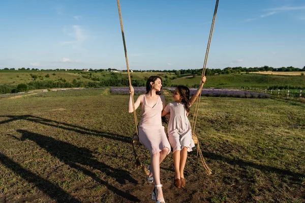 Full length of mom and daughter in pink dresses looking at each other while riding swing - foto de stock