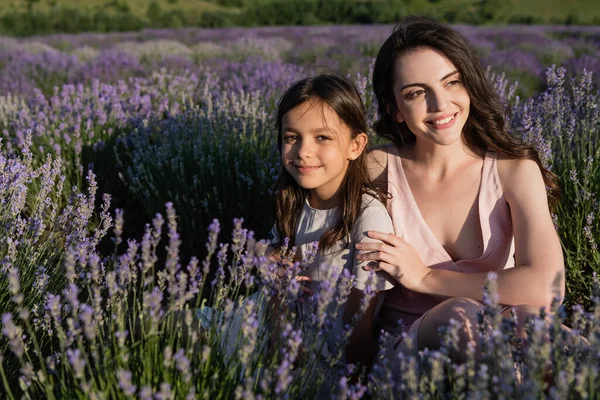 Joyful brunette mother and daughter sitting in meadow with flowering lavender — Stockfoto