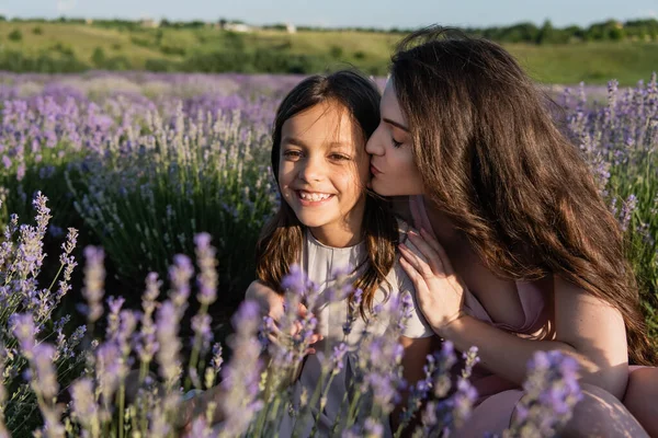 Женщина с длинными волосами целует счастливую дочь рядом с цветущей лавандой — стоковое фото