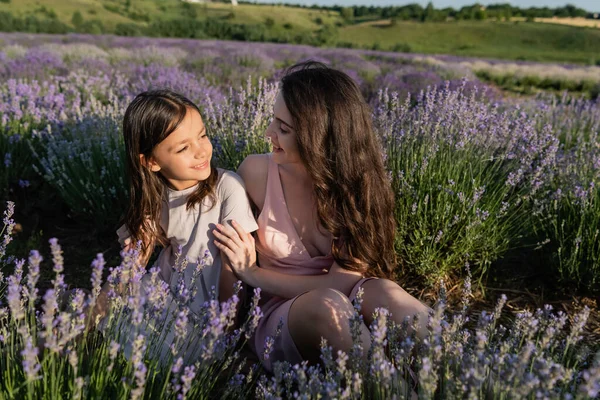 Cheerful woman and girl sitting in meadow near blooming lavender - foto de stock