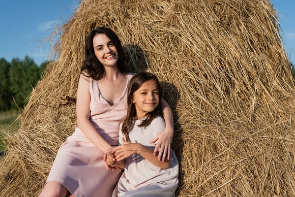 Woman and daughter in pink dresses looking at camera while sitting on haystack — Stock Photo