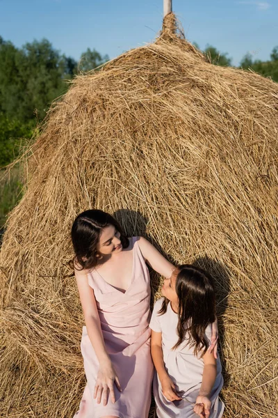 Brunette woman and girl sitting on haystack and smiling at each other — Stock Photo