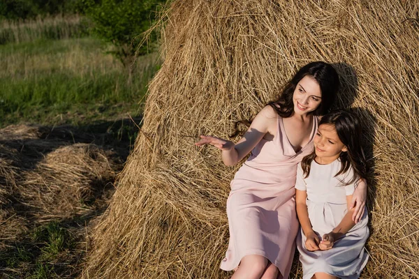Smiling woman pointing with hand while sitting on haystack with daughter — Photo de stock