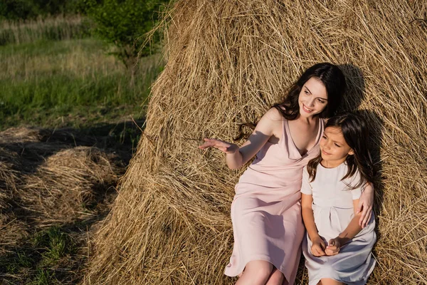 Smiling woman pointing with hand while sitting on haystack with daughter — Stock Photo