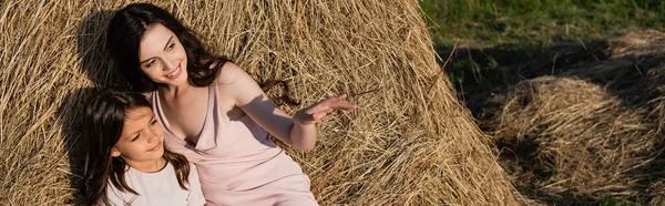 Happy woman sitting on haystack with daughter and pointing with hand, banner - foto de stock